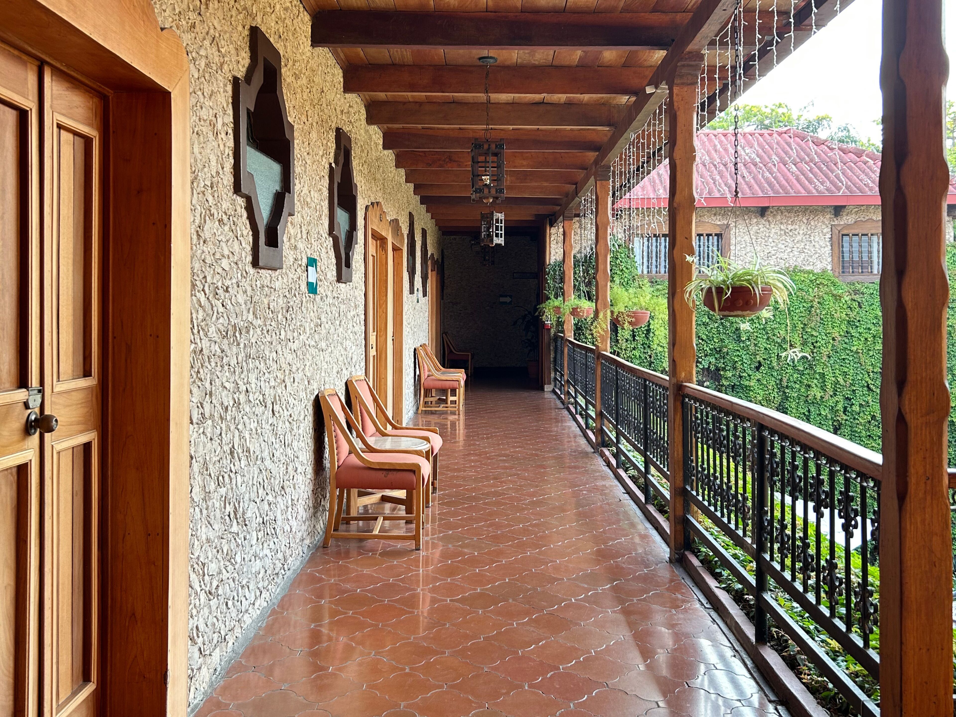 A covered porch with wooden chairs, a stone wall, and a view of a red-roofed building.