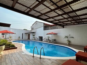 A swimming pool with a wooden deck, surrounded by lounge chairs and umbrellas, under a metal roof structure. - Hotel Del Patio (Flores)