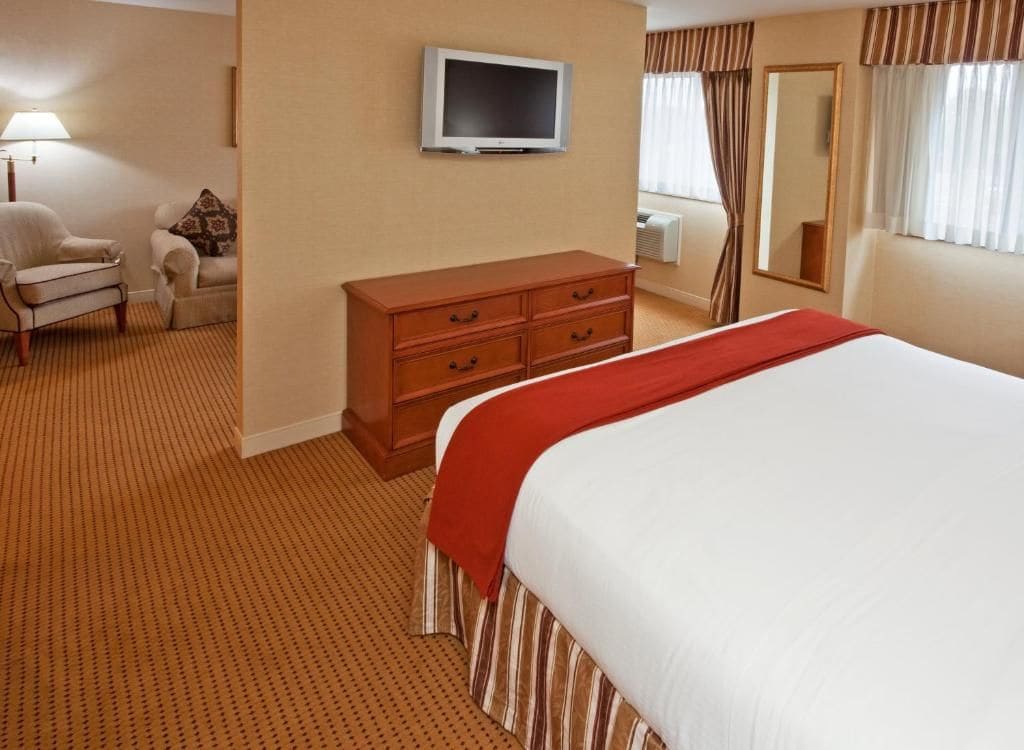 A hotel room with a large bed, a television mounted on the wall, a dresser, a chair, and a window with curtains.