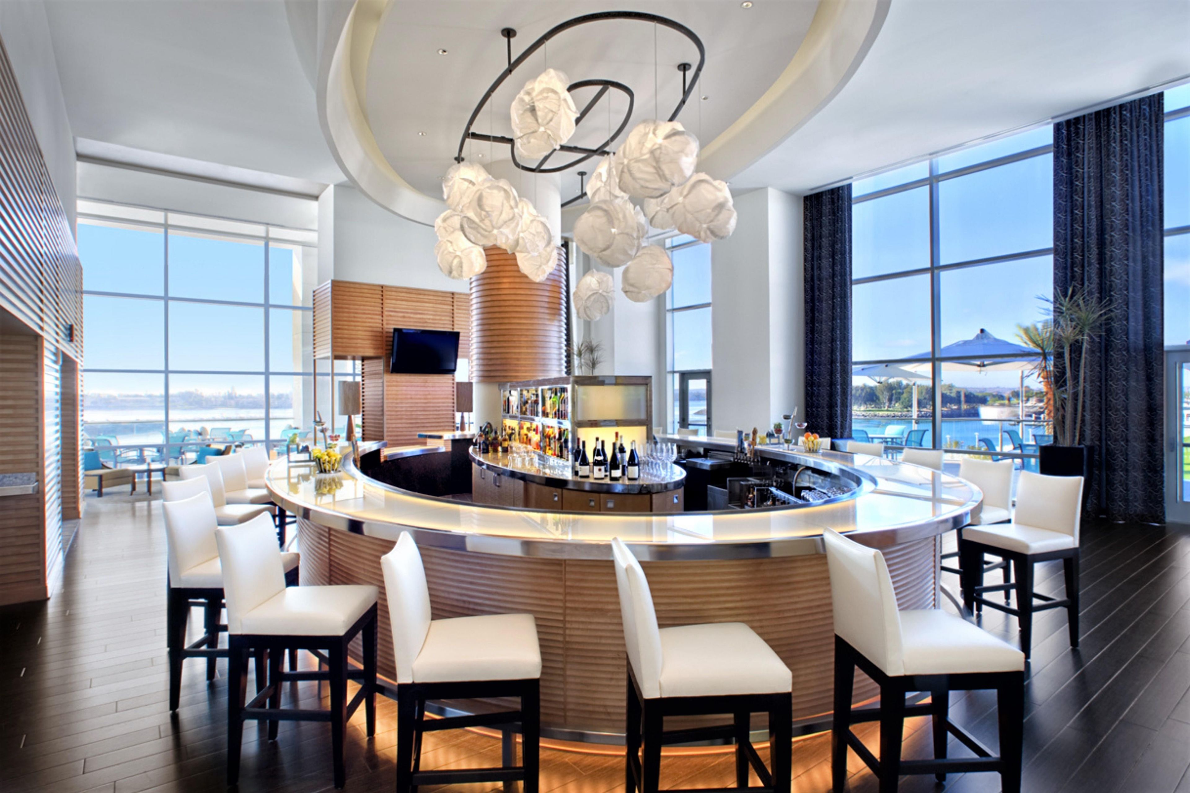 A modern bar area with a curved counter, bar stools, and a large window offering a view of the cityscape.