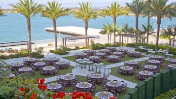 An outdoor dining area with multiple round tables and chairs, set against a backdrop of palm trees and a body of water.