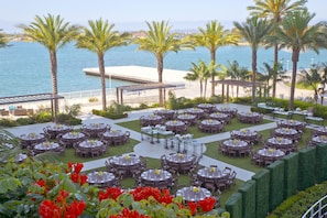 An outdoor dining area with multiple round tables and chairs, set against a backdrop of palm trees and a body of water. - Hilton San Diego Bayfront (San Diego)