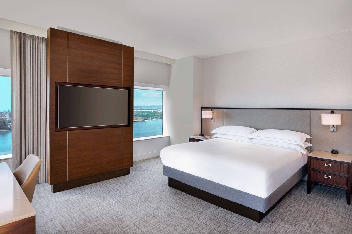 A hotel room with a large bed, a flat-screen TV mounted on a wooden panel, a desk with a chair, and a view of a body of water through a large window.