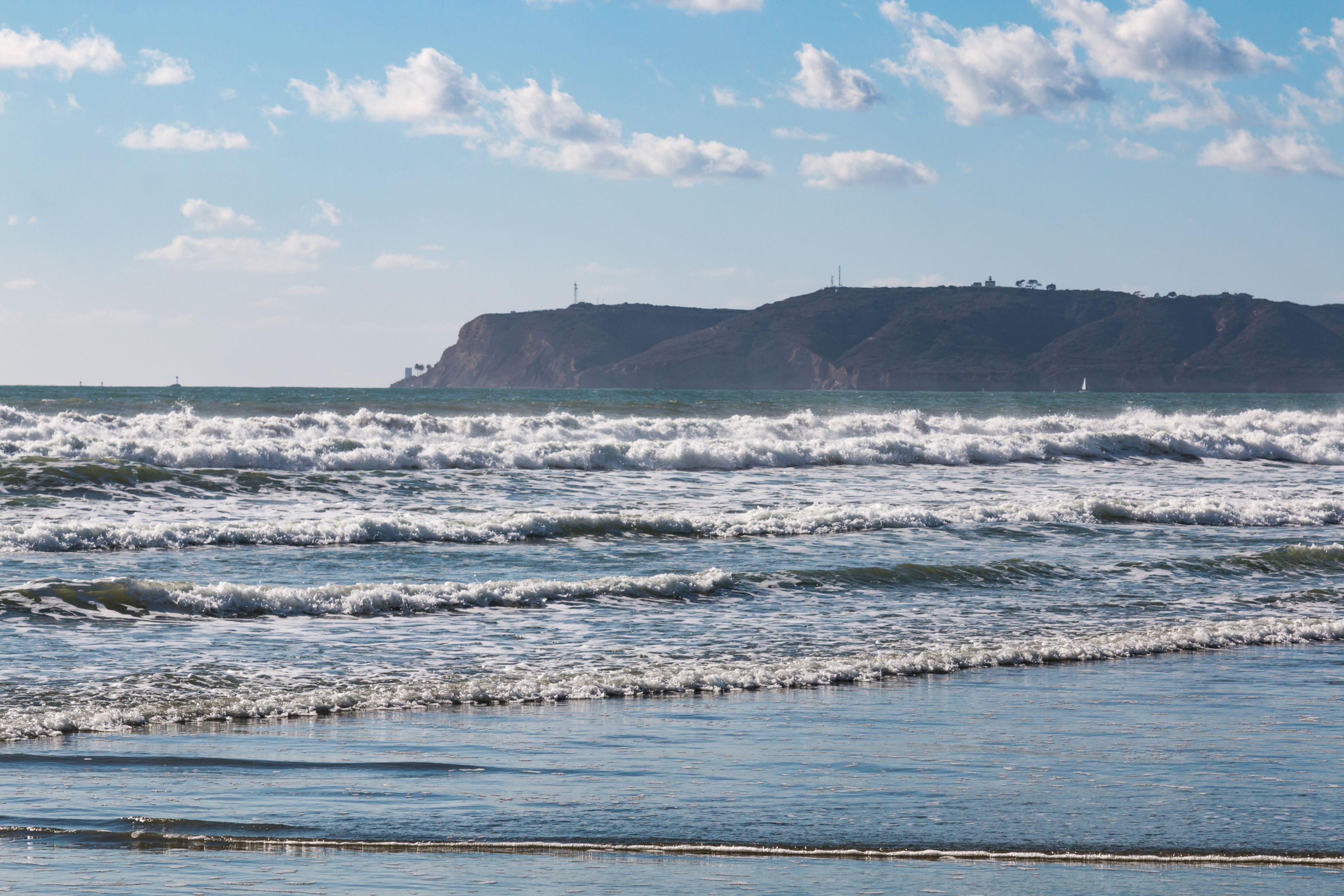 Paisaje costero con olas, cielo despejado y una colina distante con construcciones.