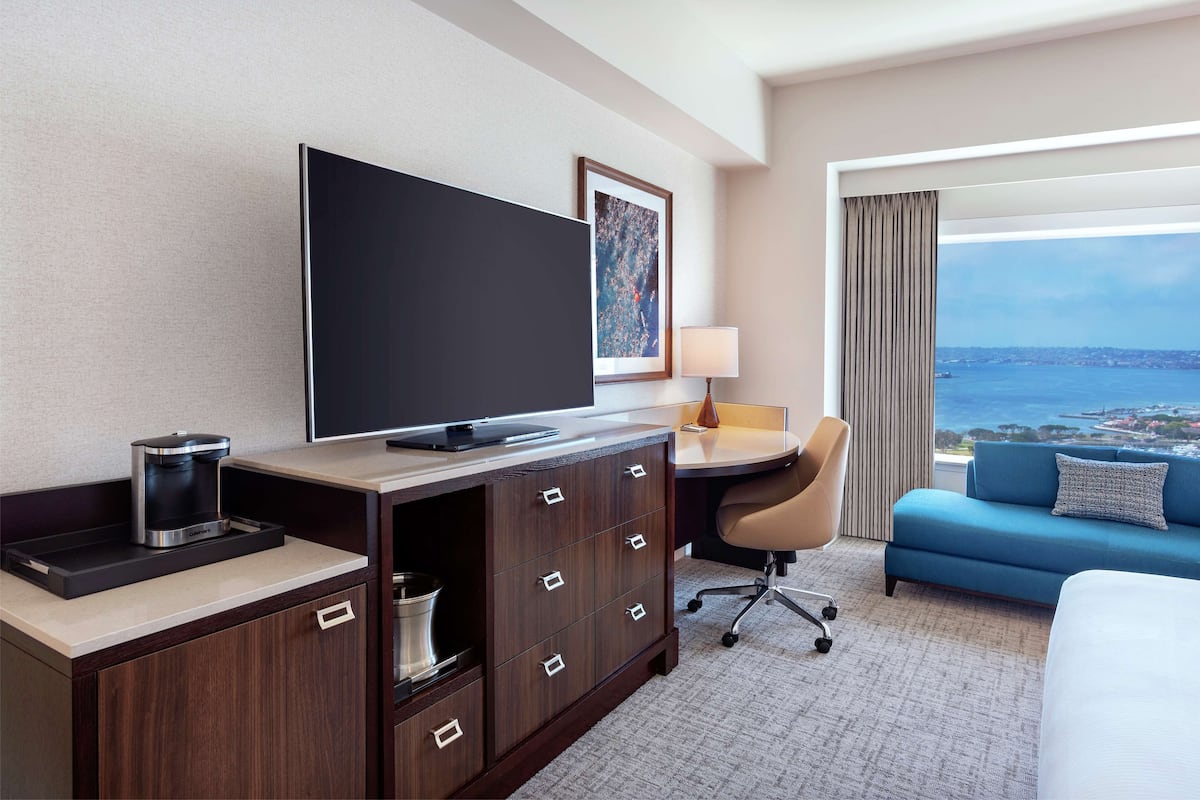 A hotel room with a bed, a desk with a chair, a television, a coffee maker, a lamp, and a view of the ocean.