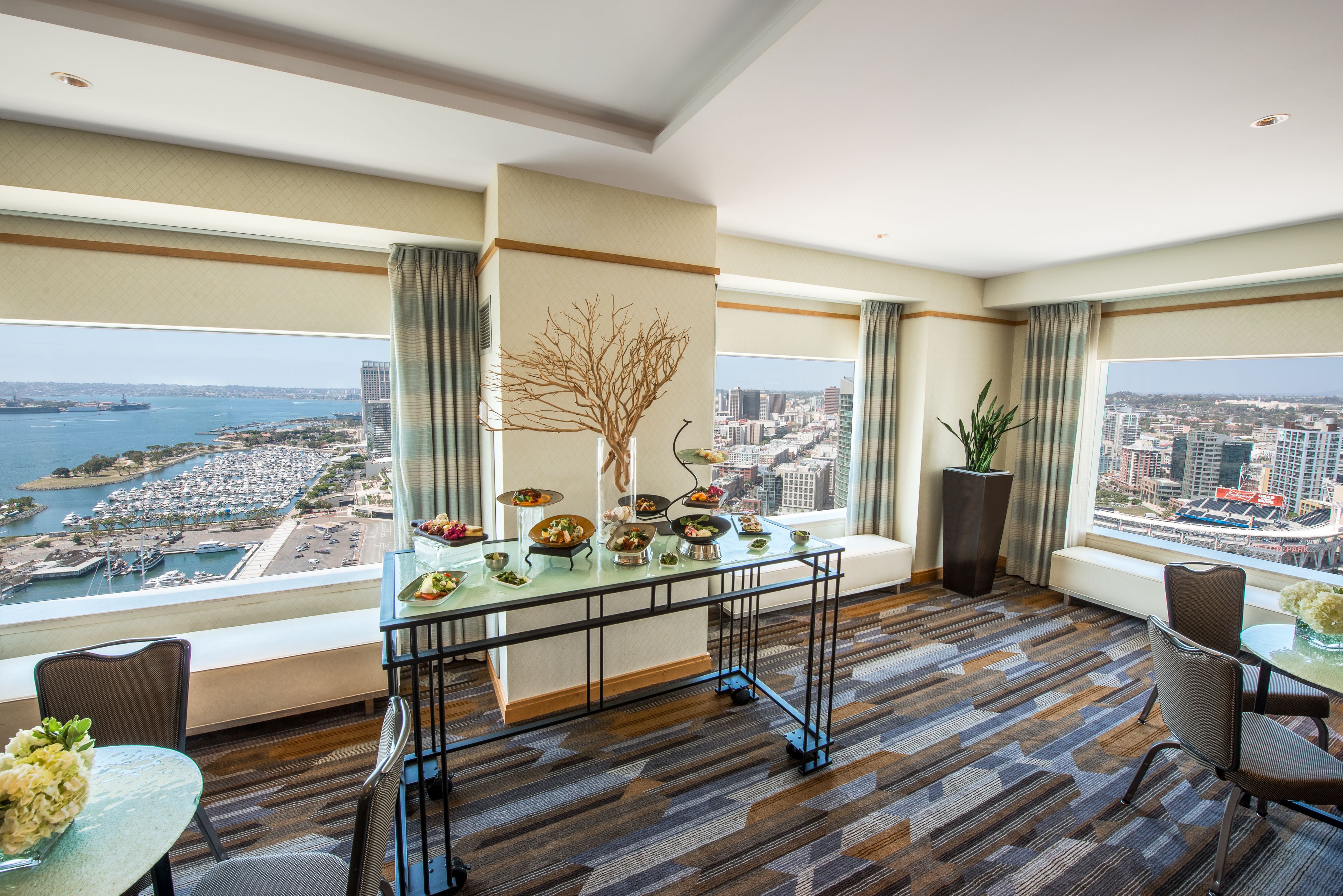A high-rise hotel room with a panoramic city and water view, a dining table with dishes, and a decorative tree sculpture.