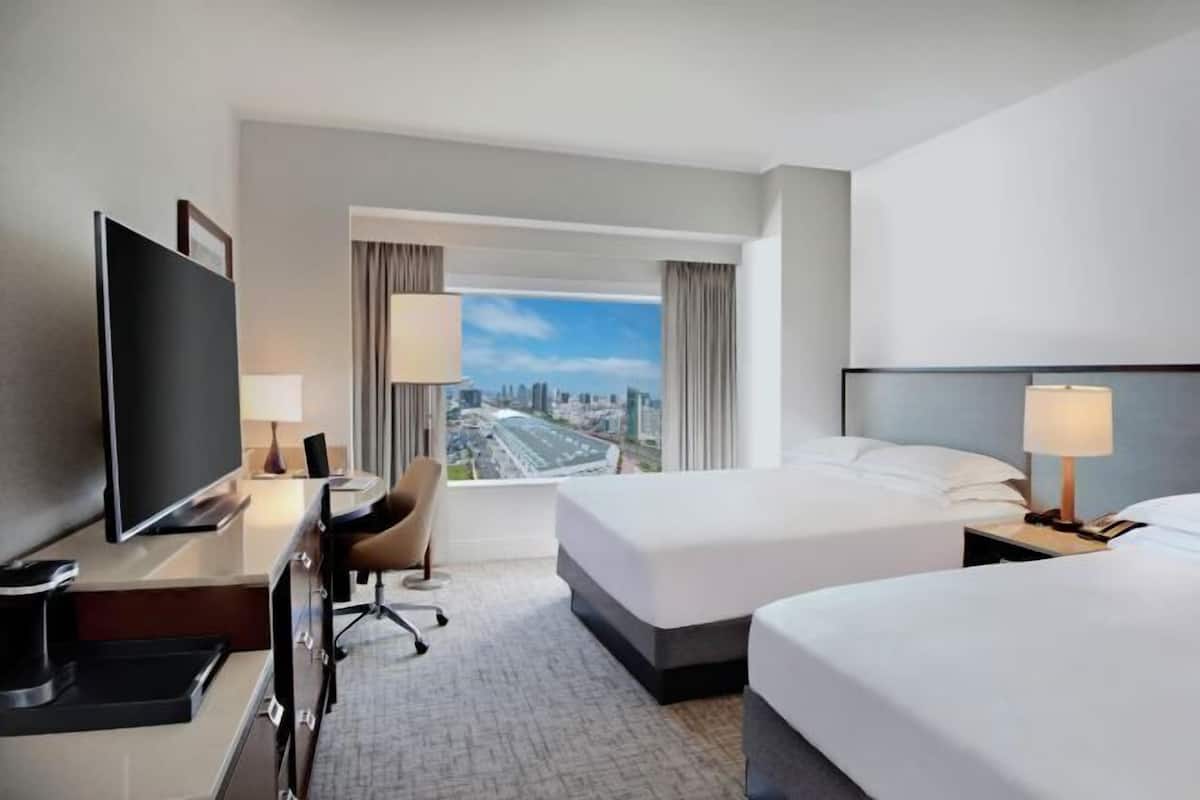 A hotel room with a large TV, two beds, a desk with a chair, and a view of a cityscape through the window.