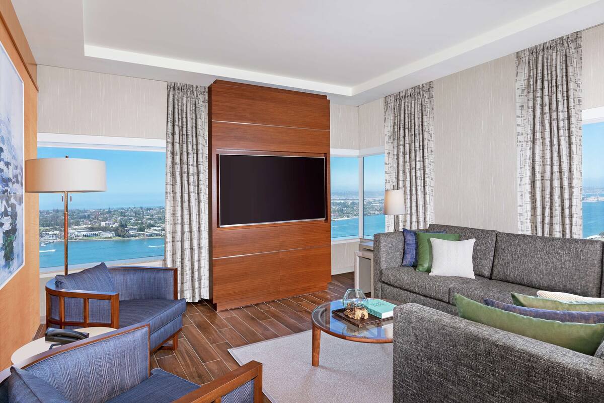 A modern living room with a large flat-screen TV, a grey sofa, a small coffee table, and a view of the city and sea through large windows.