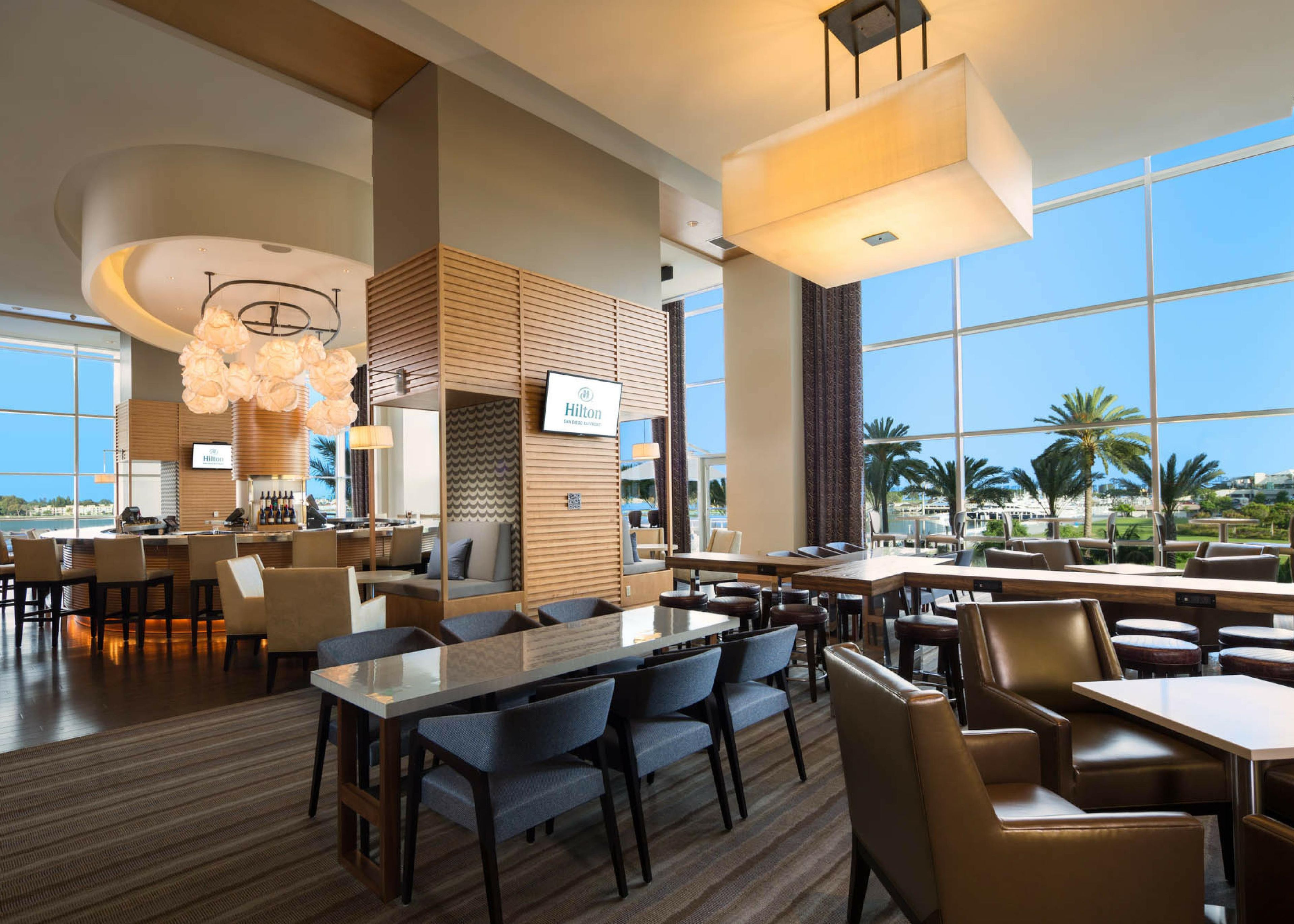 A spacious hotel lobby with a bar area, seating arrangements, and large windows offering a view of palm trees and a pool.