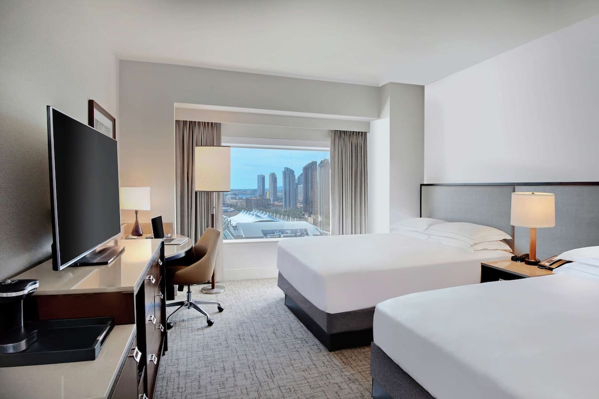A hotel room with a large bed, a desk with a TV, a chair, and a view of a cityscape.