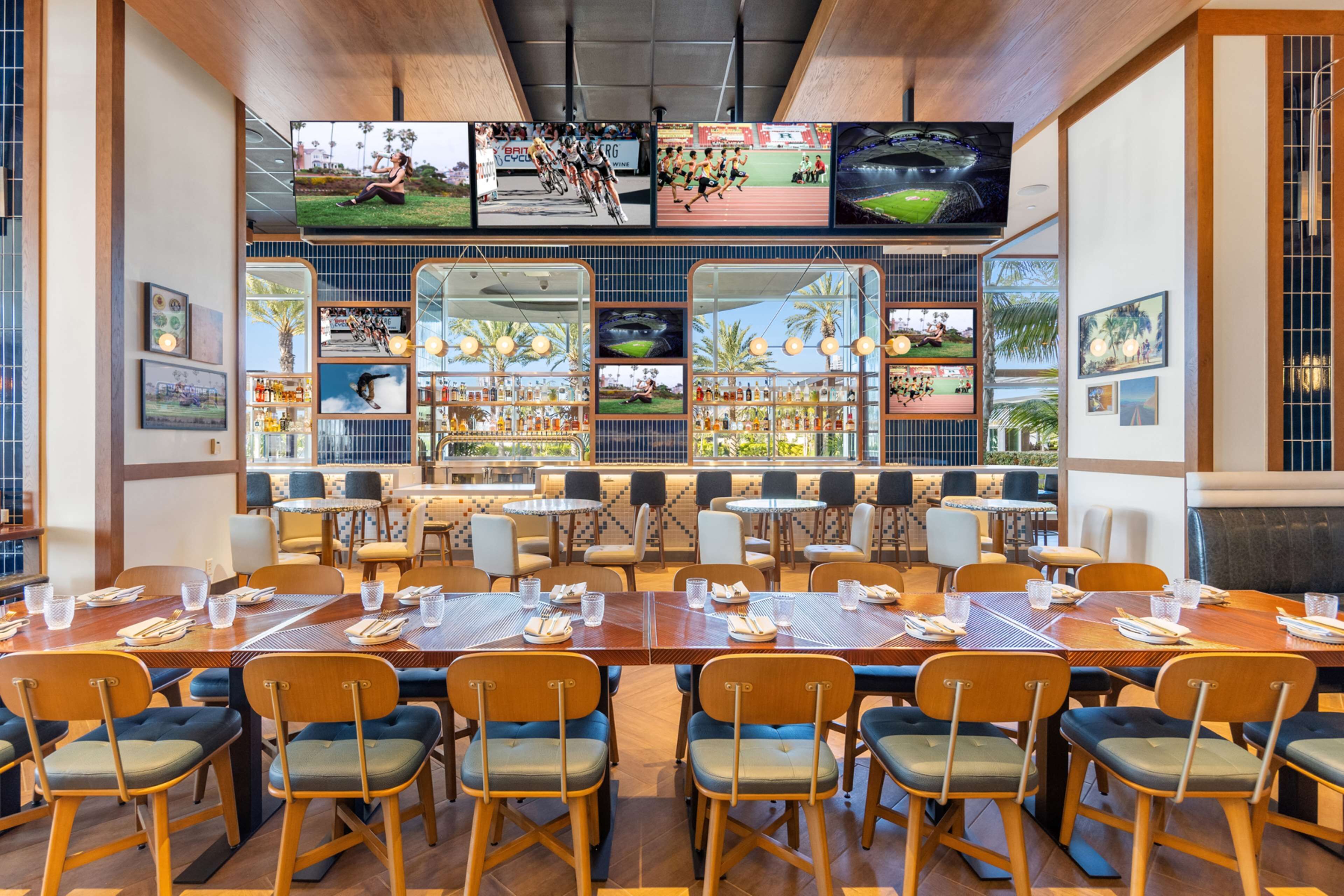 A restaurant with multiple dining tables and chairs, a large TV screen displaying sports, and a view of palm trees through the windows.