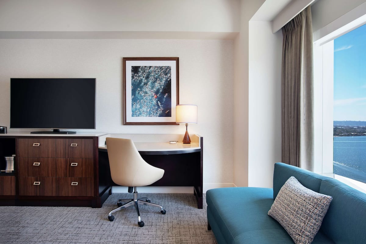 A hotel room with a desk, chair, TV, and a sofa with a view of the sea.