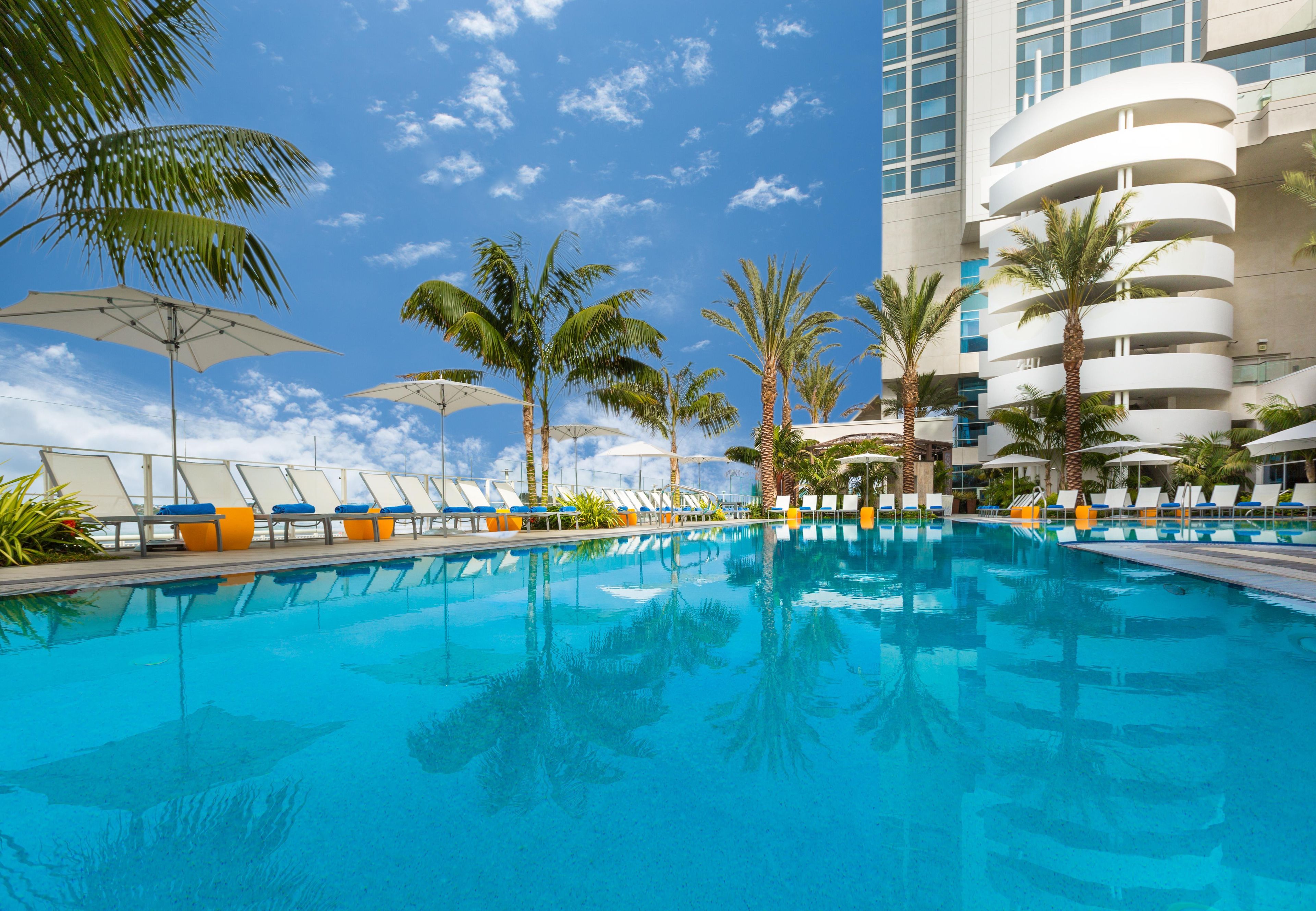 A swimming pool with lounge chairs and umbrellas, surrounded by palm trees and a modern building.