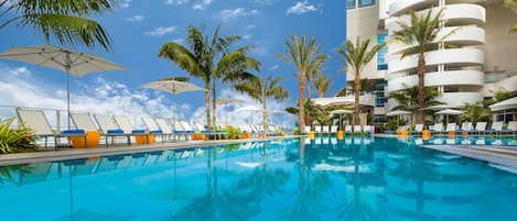 Una piscina con sillas de descanso y sombrillas, rodeada de palmeras y un edificio moderno.