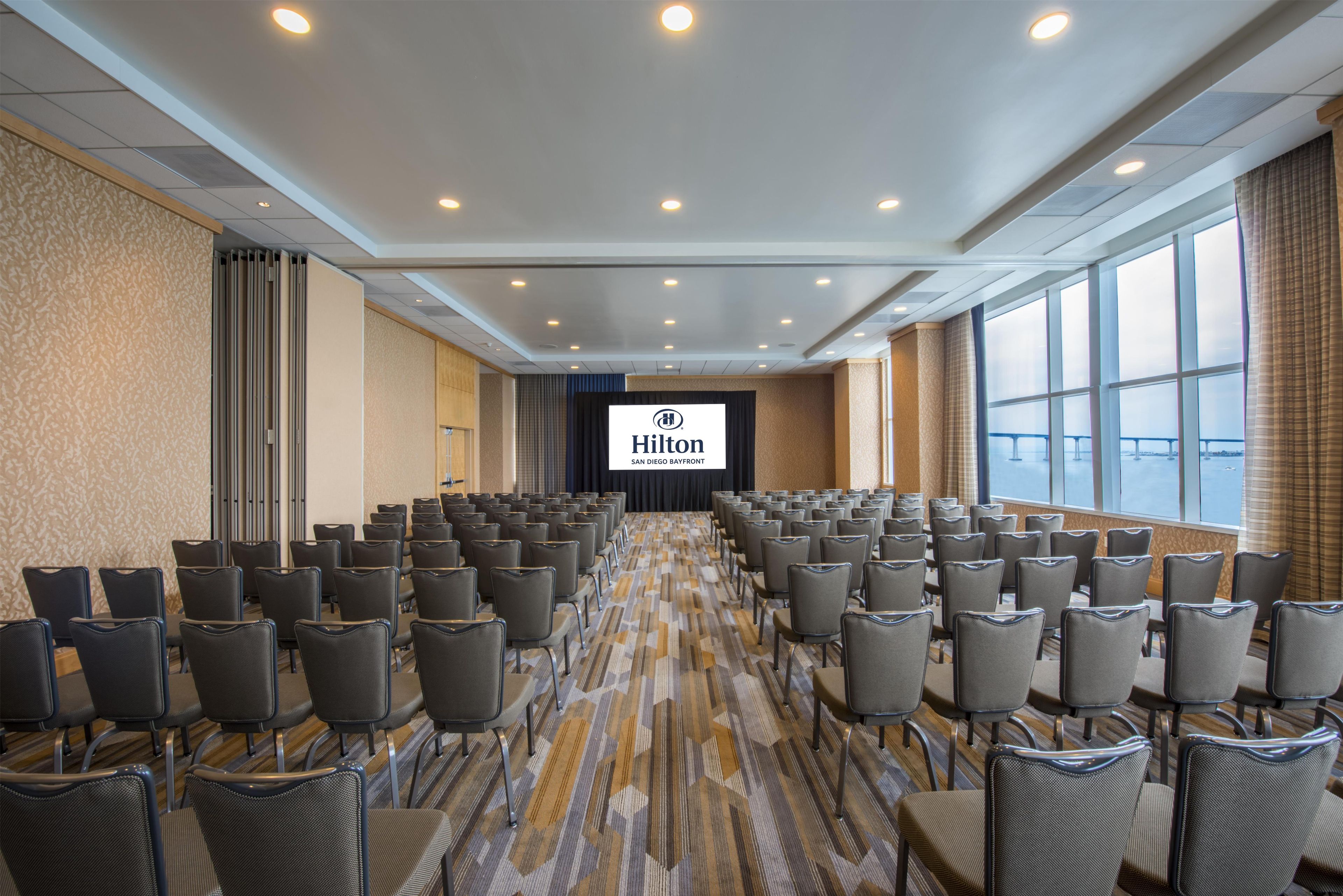 A large conference room with rows of chairs, a stage with a screen displaying the Hilton logo, and large windows offering a view.