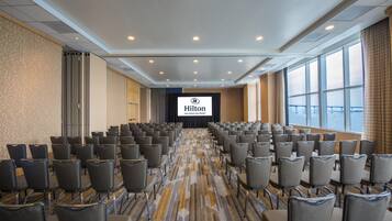A large conference room with rows of chairs, a stage with a screen displaying the Hilton logo, and large windows offering a view.