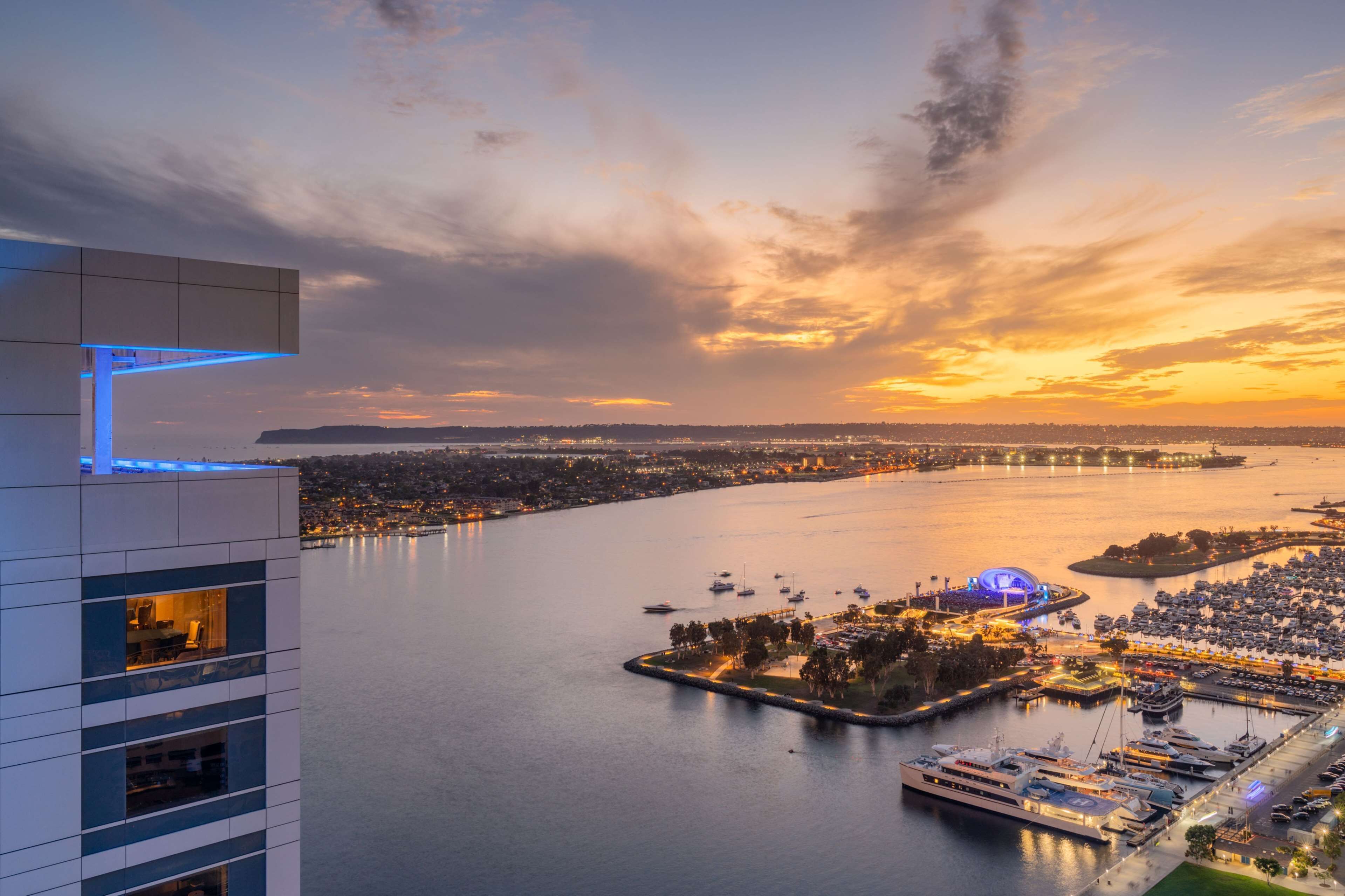 Vista para a frente marítima com um porto de iates, um edifício grande com varandas e um pôr do sol sobre a água.