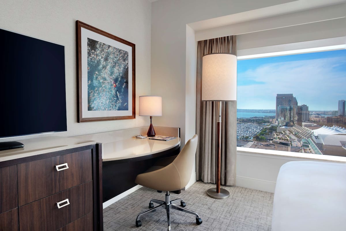 A hotel room with a desk, chair, TV, lamp, and a city view through the window.