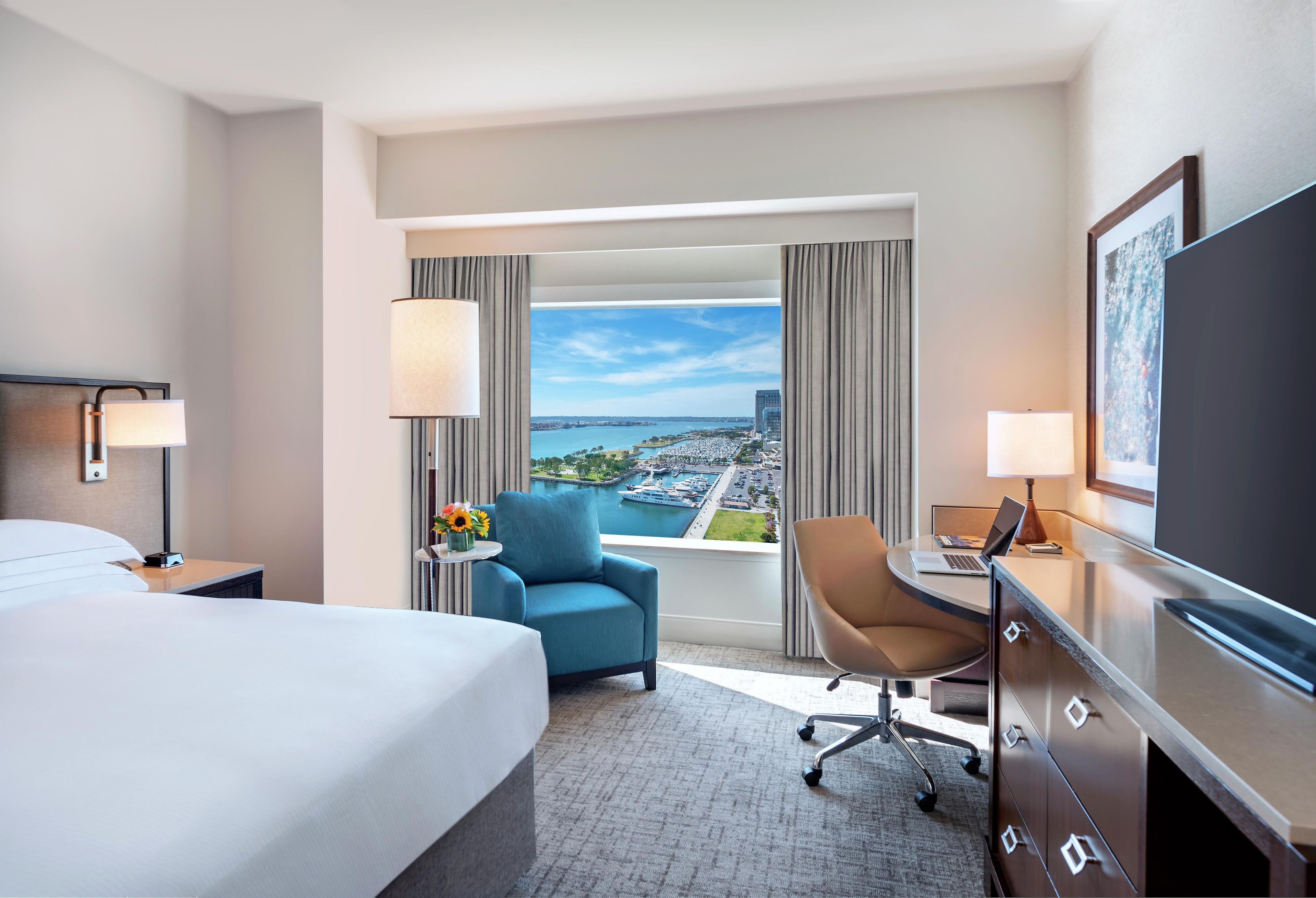 Habitación de hotel con cama, escritorio, silla, televisión y vistas a la ciudad y al agua.