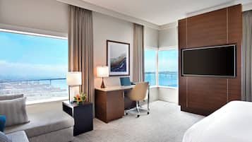 Habitación de hotel con una cama grande, un escritorio con una computadora portátil, una silla, un televisor y vistas a un puente y al agua.