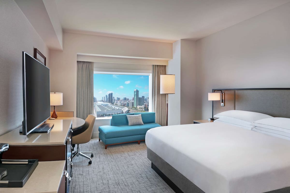A hotel room with a large bed, a desk with a flat-screen TV, a blue sofa, and a city view.