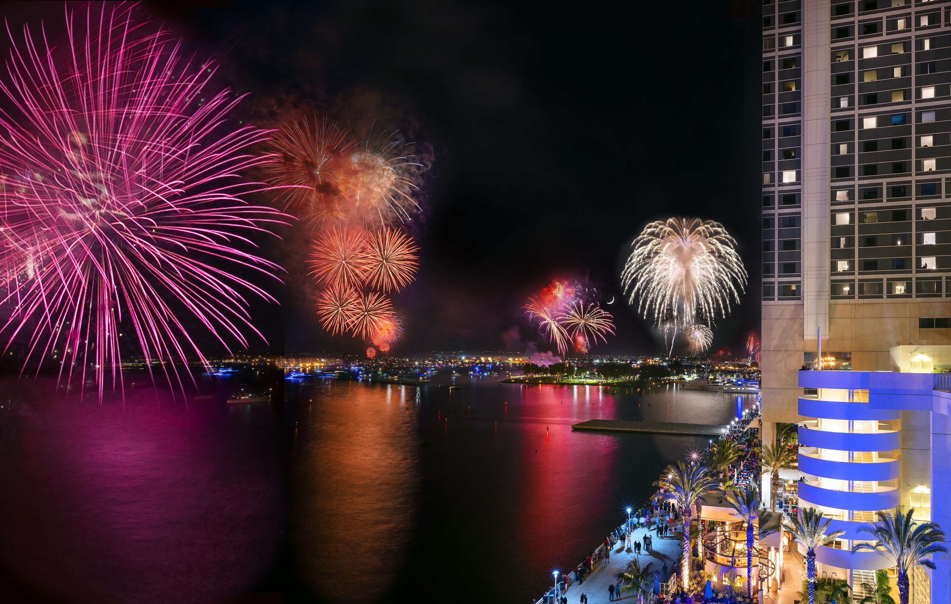 Une vue nocturne d’un front de mer illuminé par un feu d’artifice, d’un bâtiment moderne doté de balcons et d’une promenade bien éclairée.