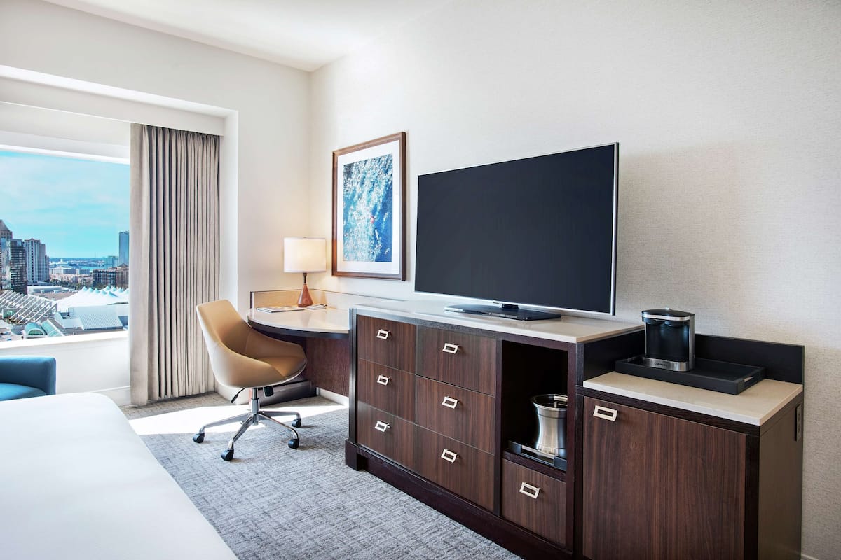 A hotel room with a large bed, a desk with a chair, a television, a coffee maker, and a view of the city.