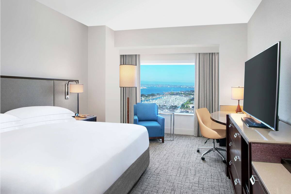 A hotel room with a large bed, a desk with a flat-screen TV, a chair, a desk lamp, and a view of the city and sea through the window.