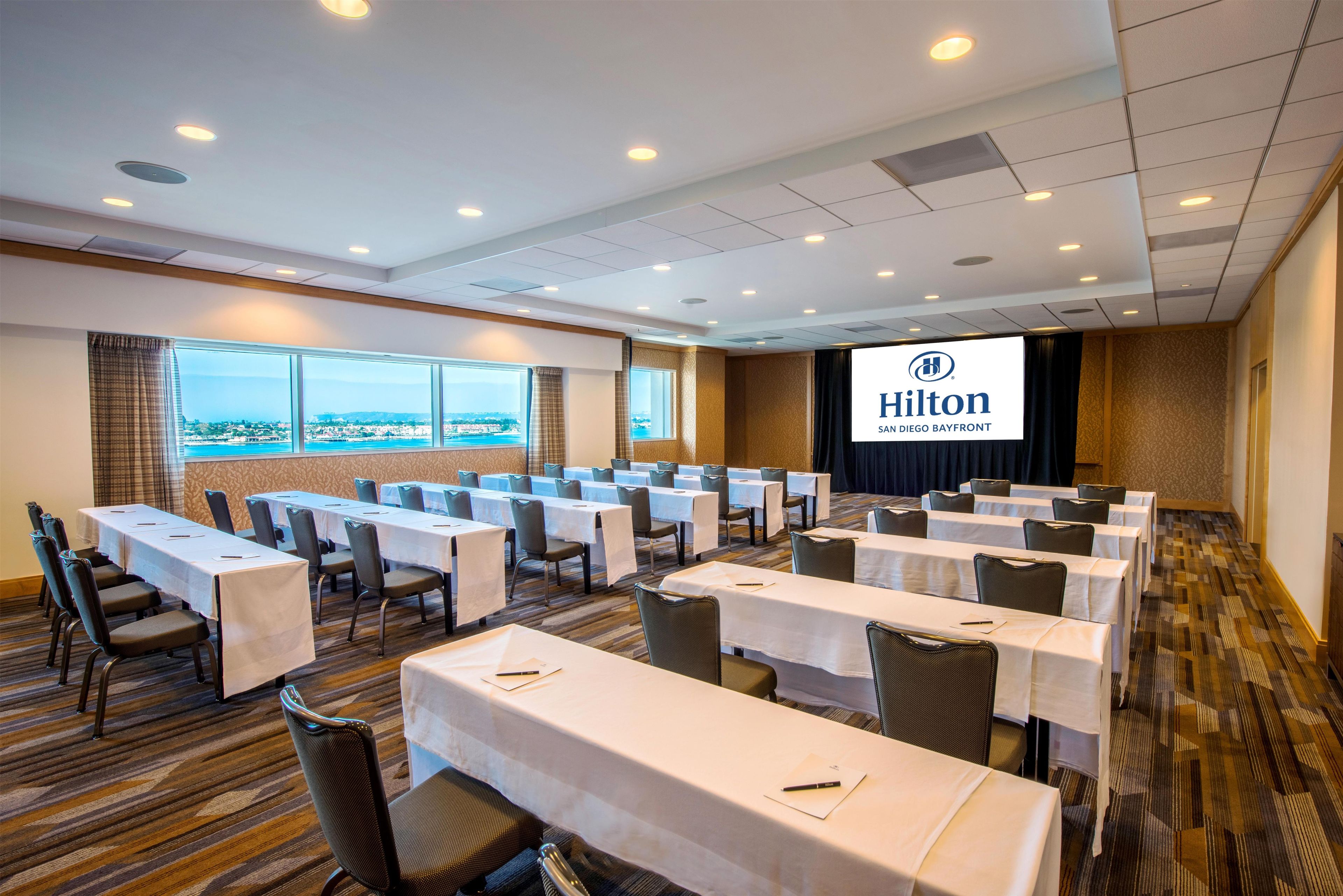 A conference room with multiple tables and chairs, a large screen displaying the Hilton logo, and a view of the city through large windows.