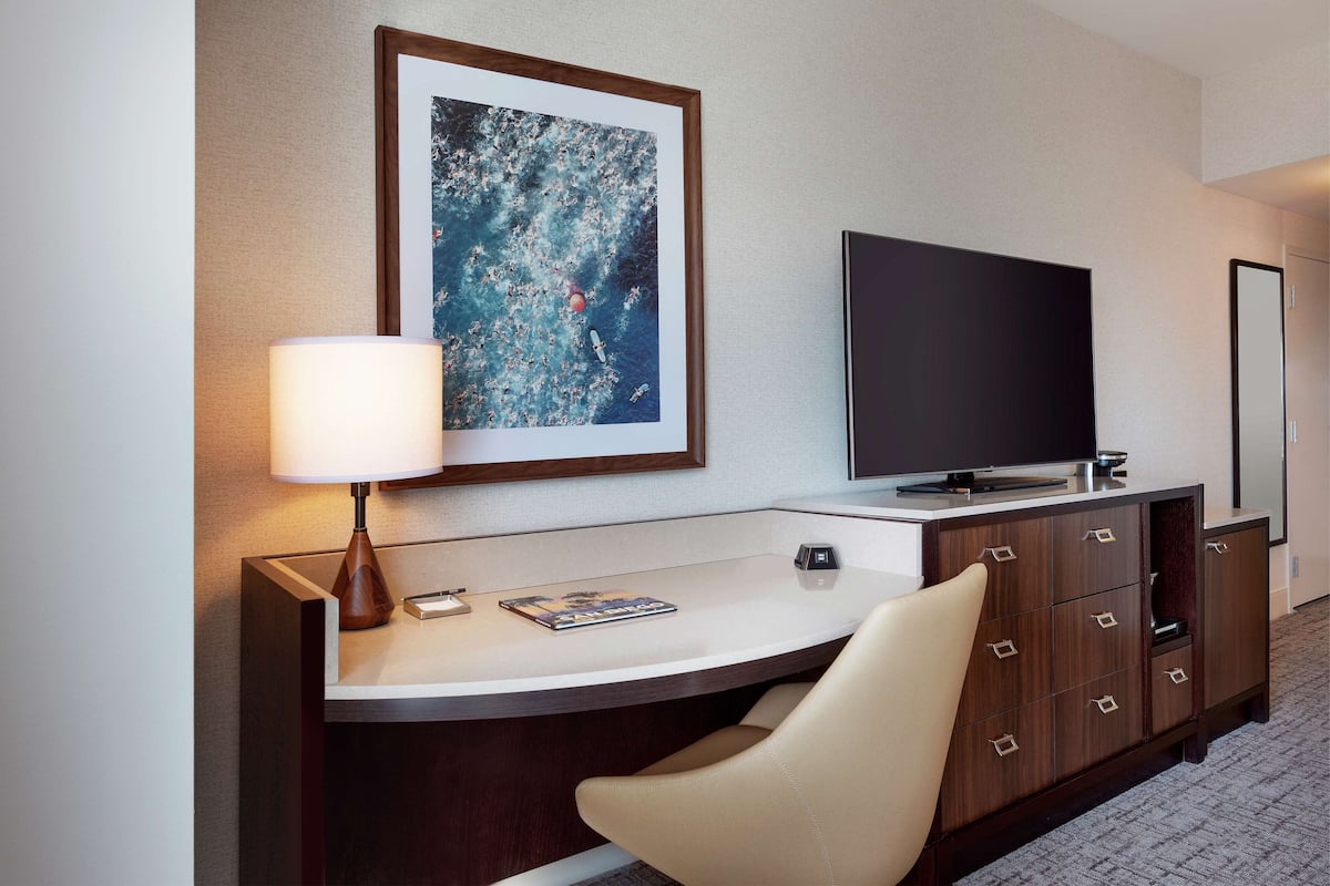 A hotel room with a desk, a television, a lamp, a framed picture, and a mirror.