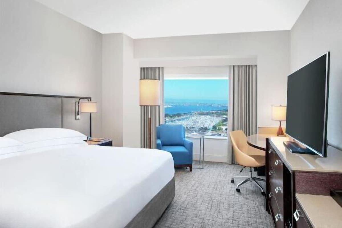 Habitación de hotel con cama, escritorio, silla, televisor y vista a la ciudad.