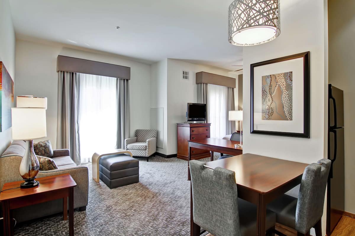 A hotel room with a sofa, ottoman, dining table, chairs, a TV, a desk, a lamp, a framed picture, and a patterned carpet.