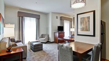 A hotel room with a sofa, ottoman, dining table, chairs, a TV, a desk, a lamp, a framed picture, and a patterned carpet.