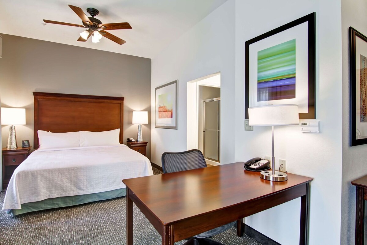 Habitación de hotel con cama, escritorio, silla, teléfono, lámpara, ventilador de techo y cuadros en la pared.