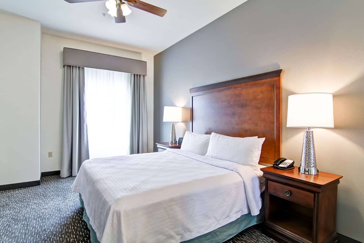 A hotel room with a bed, two bedside lamps, a telephone, a nightstand, a ceiling fan, and a window with curtains.