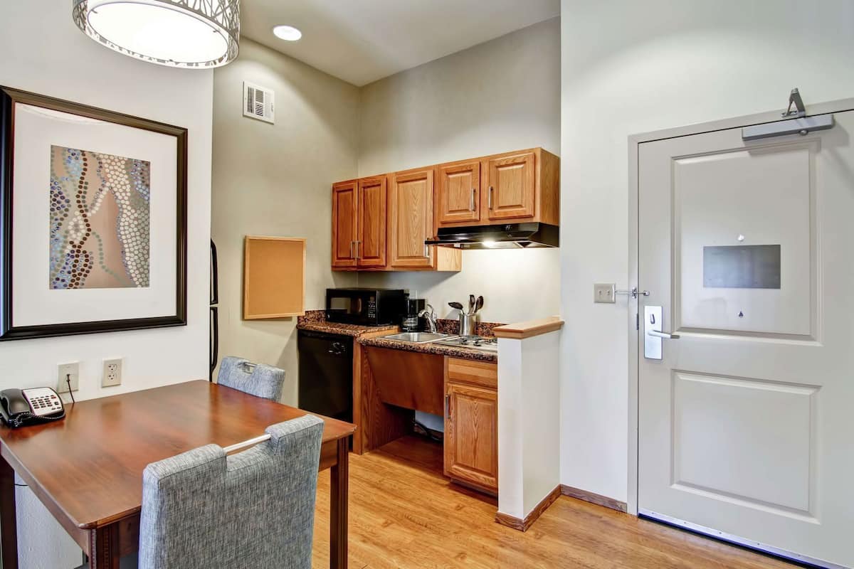 A compact kitchen with wooden cabinets, a microwave, and a sink. There's a dining table with chairs and a framed picture on the wall.