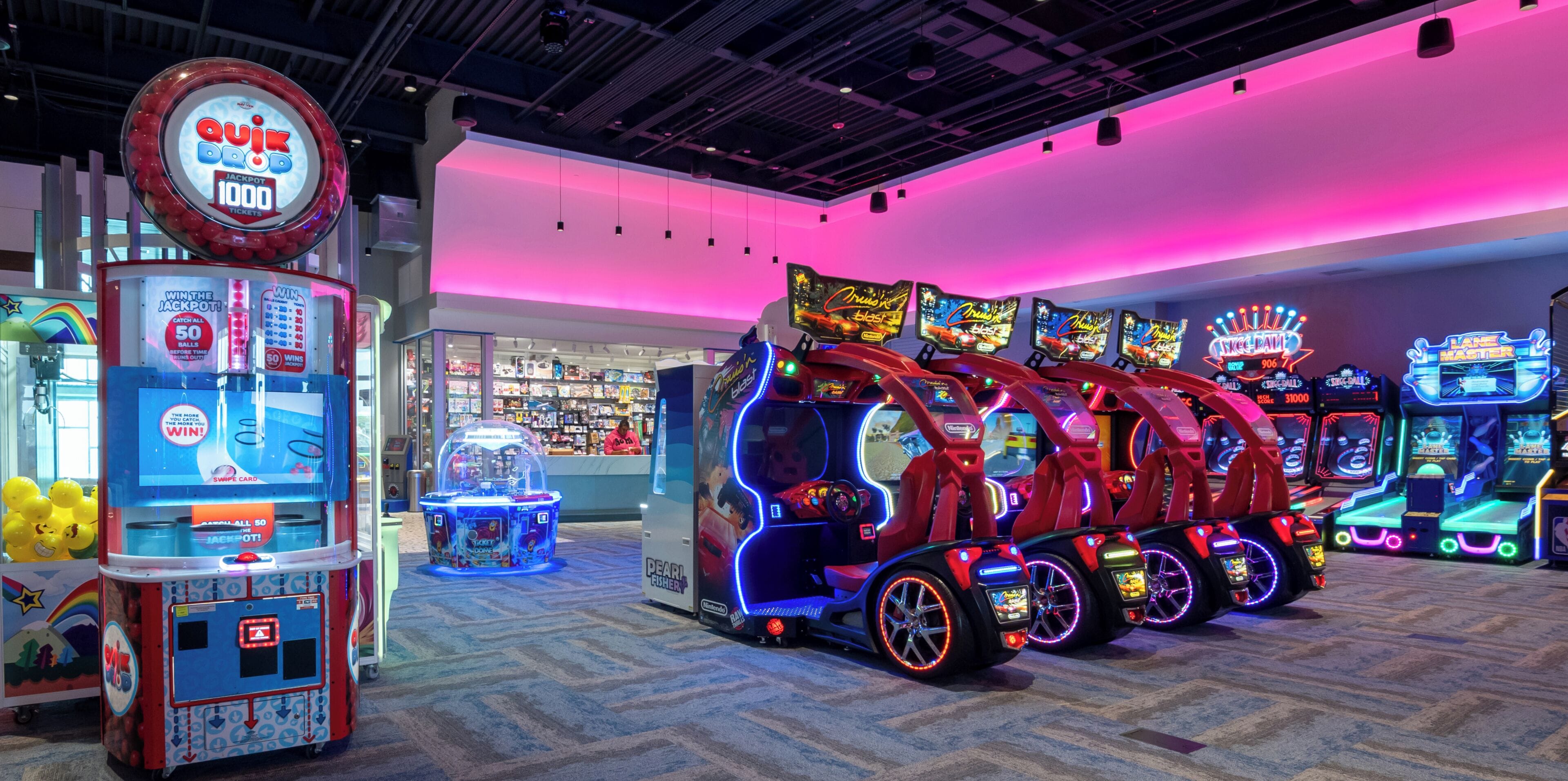 Un salón de juegos bien iluminado con diversas máquinas, incluyendo una máquina de premios 'Quik Draw' y un juego de carreras 'Pearl Fusher'.