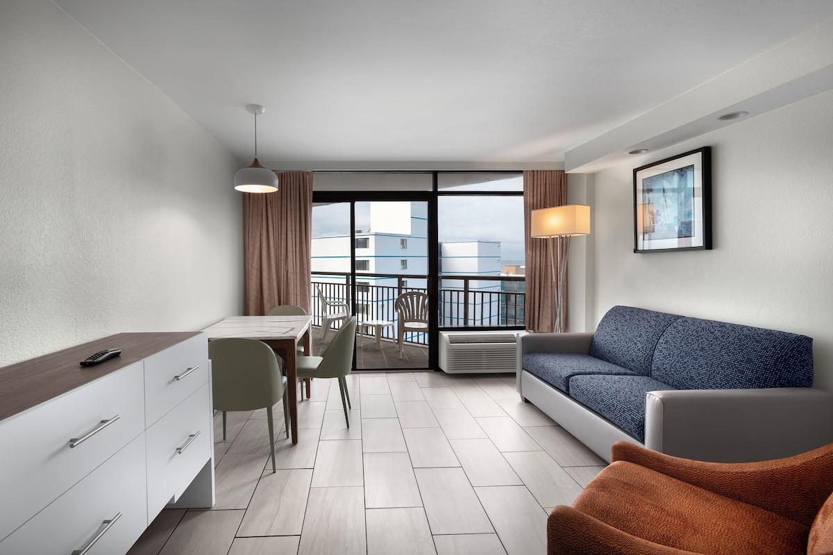 A modern hotel room with a dining area, a sofa, and a balcony with a view of buildings.