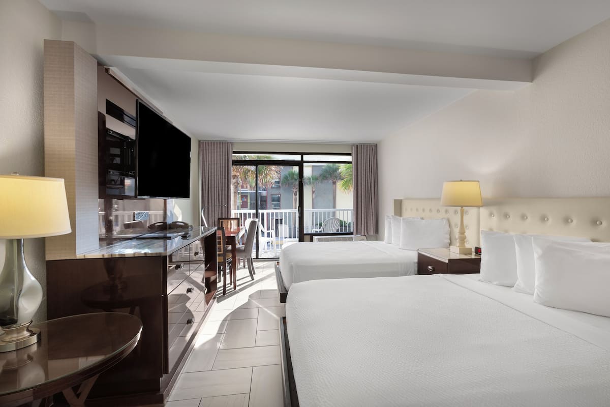 A hotel room with two beds, a desk with a TV, a dining area, and a balcony with a view of palm trees.