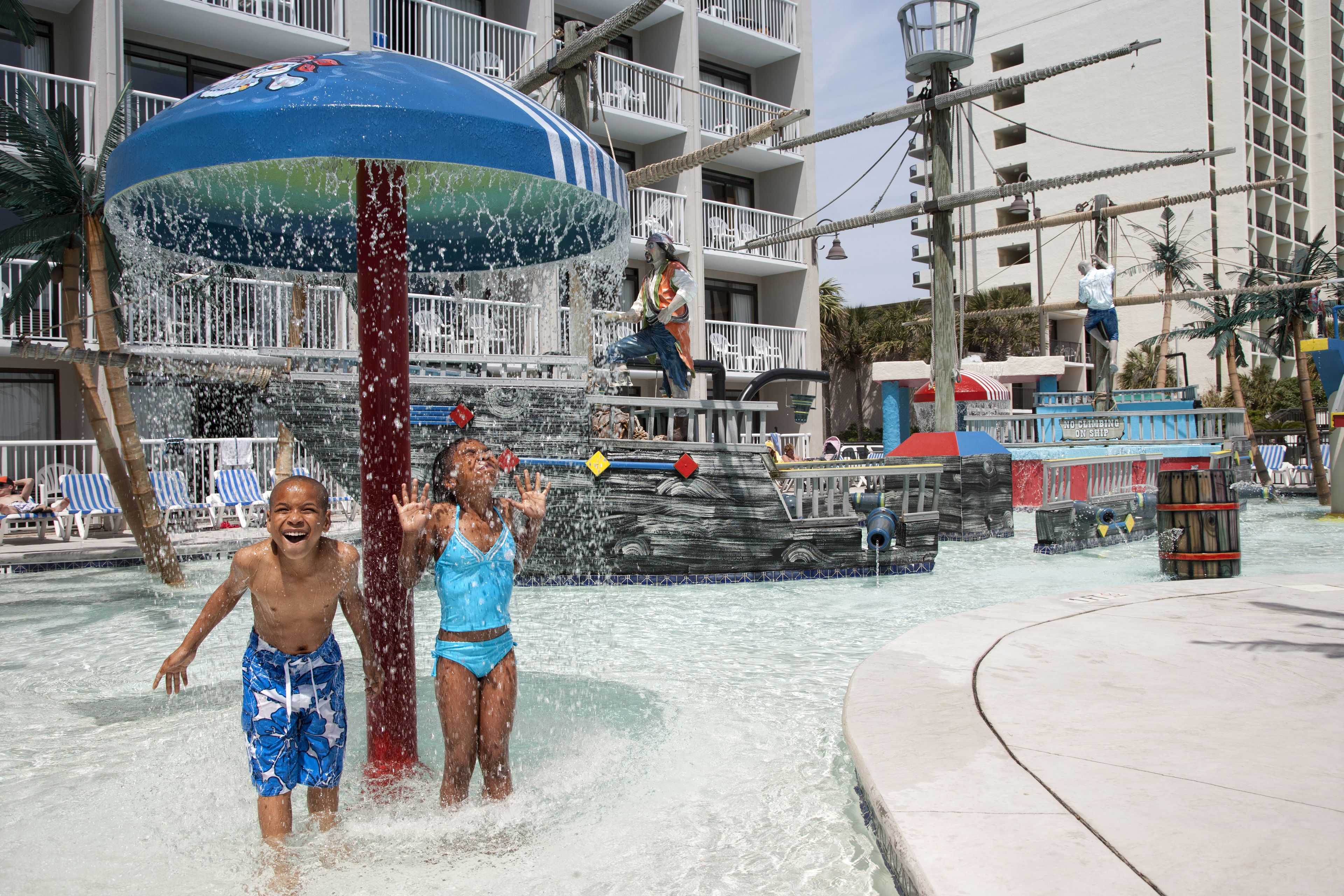 Een waterpark met een grote waterfontein, een zwembad en een gebouw met balkons op de achtergrond.