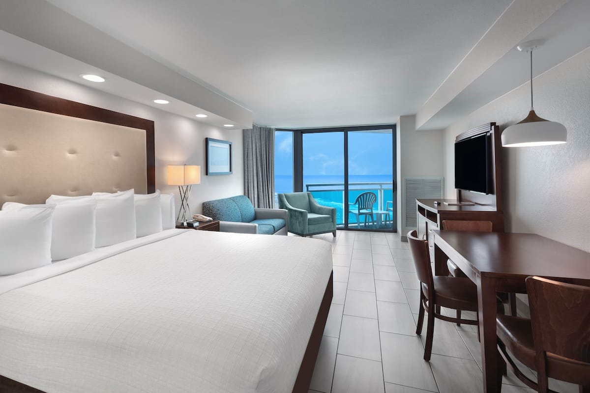 A hotel room with a large bed, a desk, a chair, a TV, and a balcony with a view of the ocean.