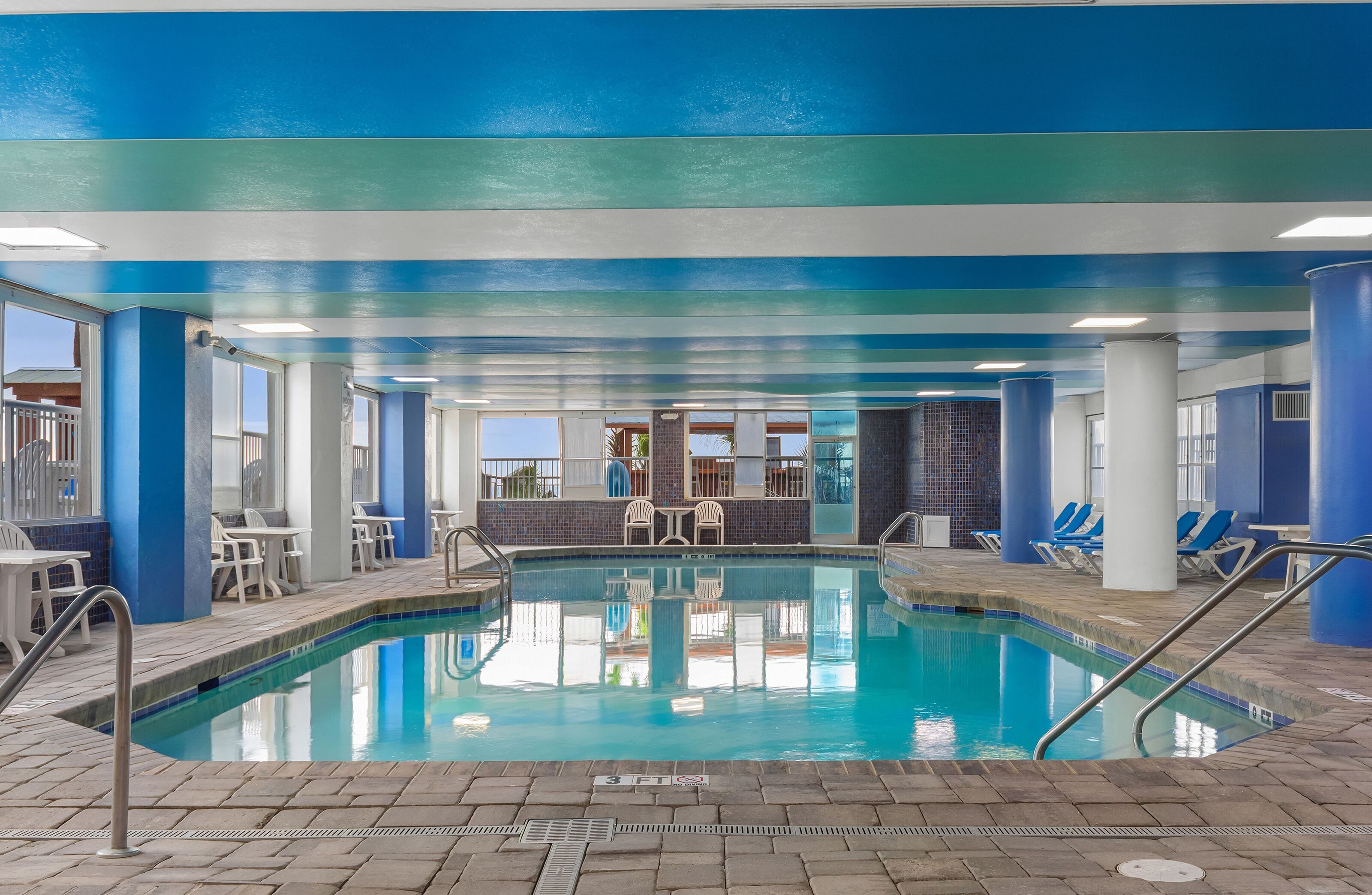 Una piscina interior con paredes azules y blancas, suelo de baldosas y sillones de descanso alrededor.