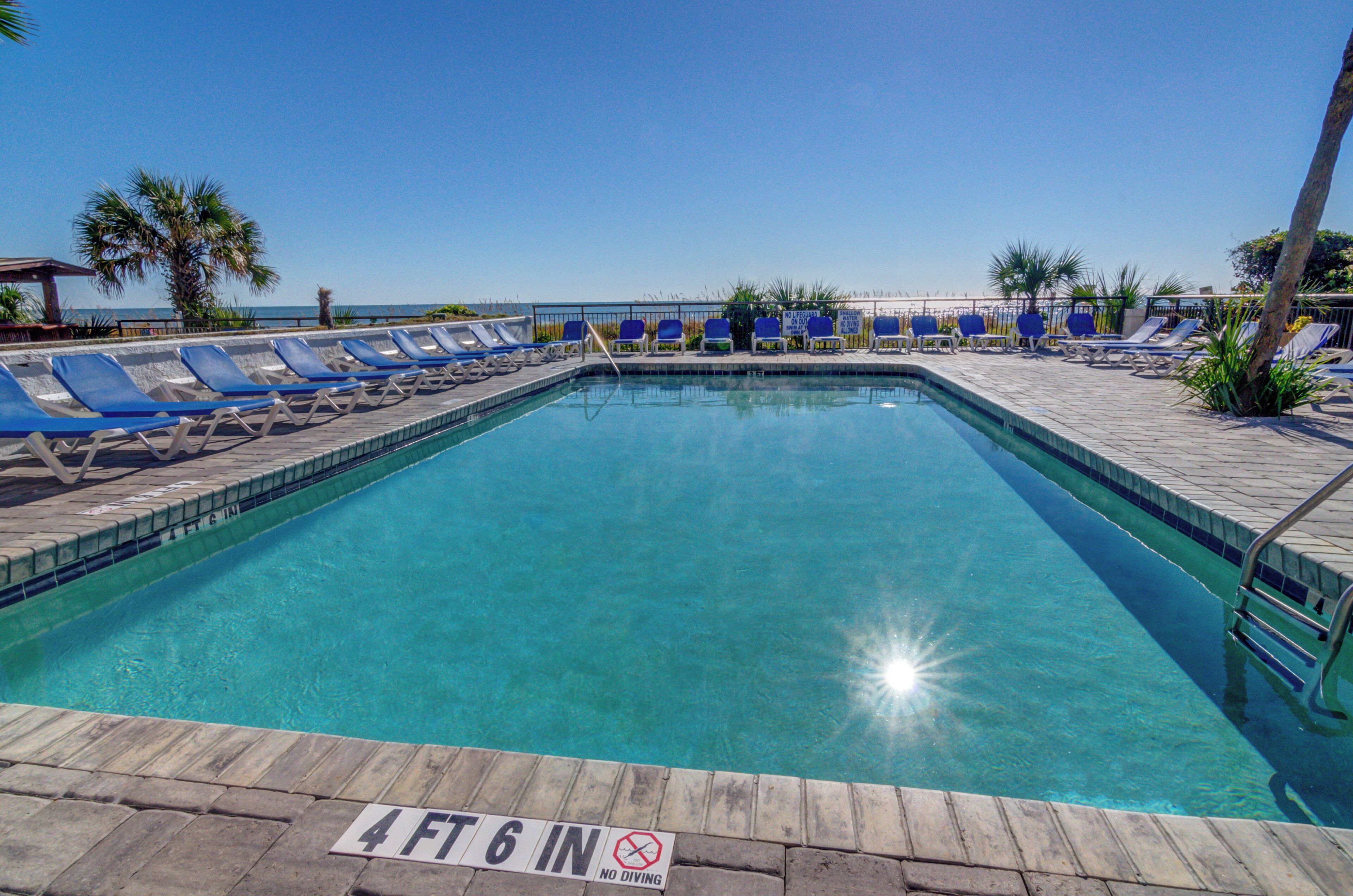 Piscina rectangular con cubierta de madera y sillas de descanso azules. Un cartel indica una profundidad de 1,4 metros con el símbolo de prohibición de saltos.