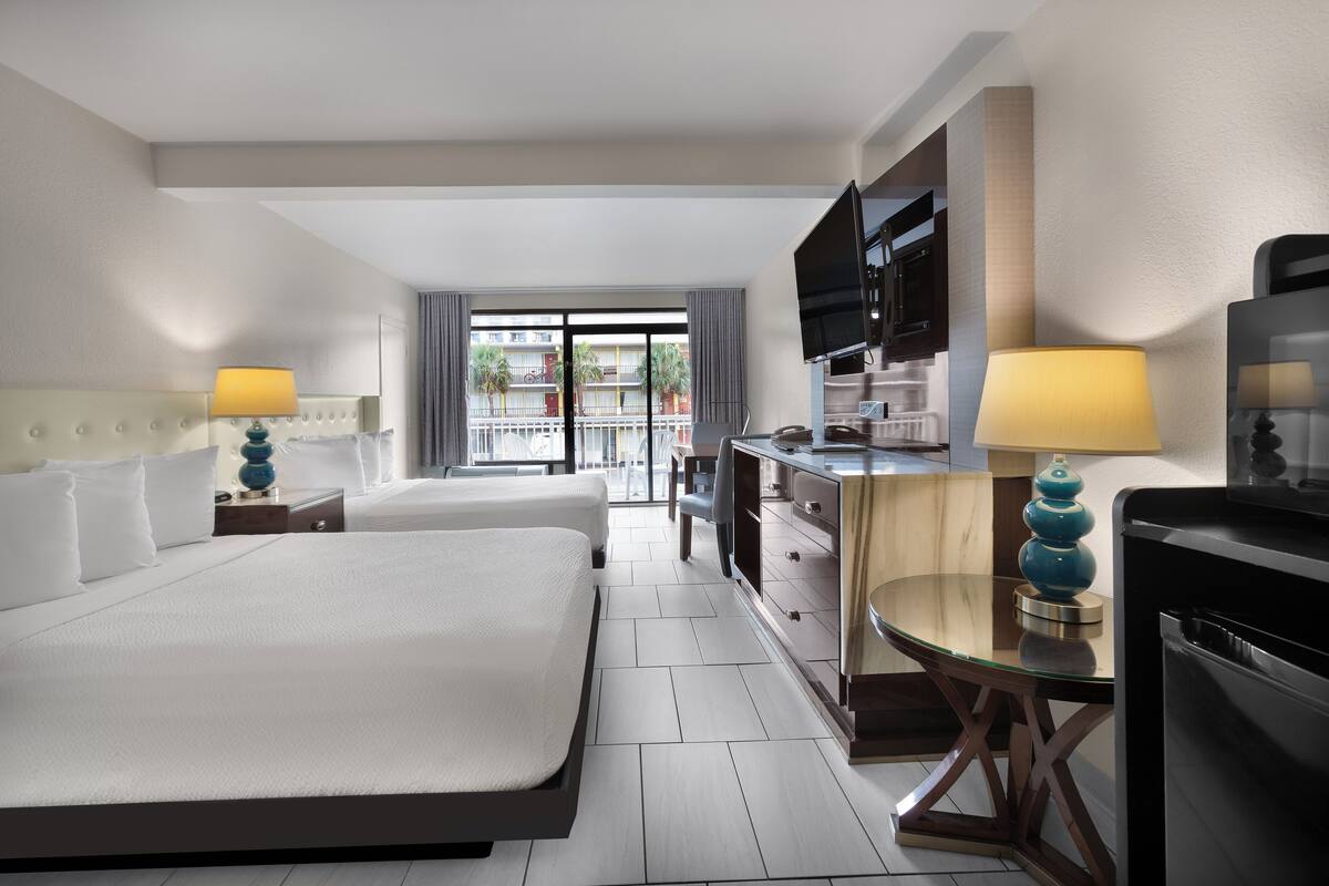 A hotel room with two beds, a desk, a TV, and a balcony with a view of buildings.