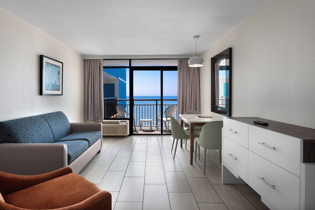 A modern hotel room with a balcony, a sofa, a dining table, and a kitchenette.