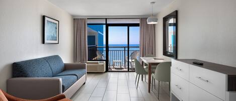 A modern hotel room with a balcony, a sofa, a dining table, and a kitchenette.