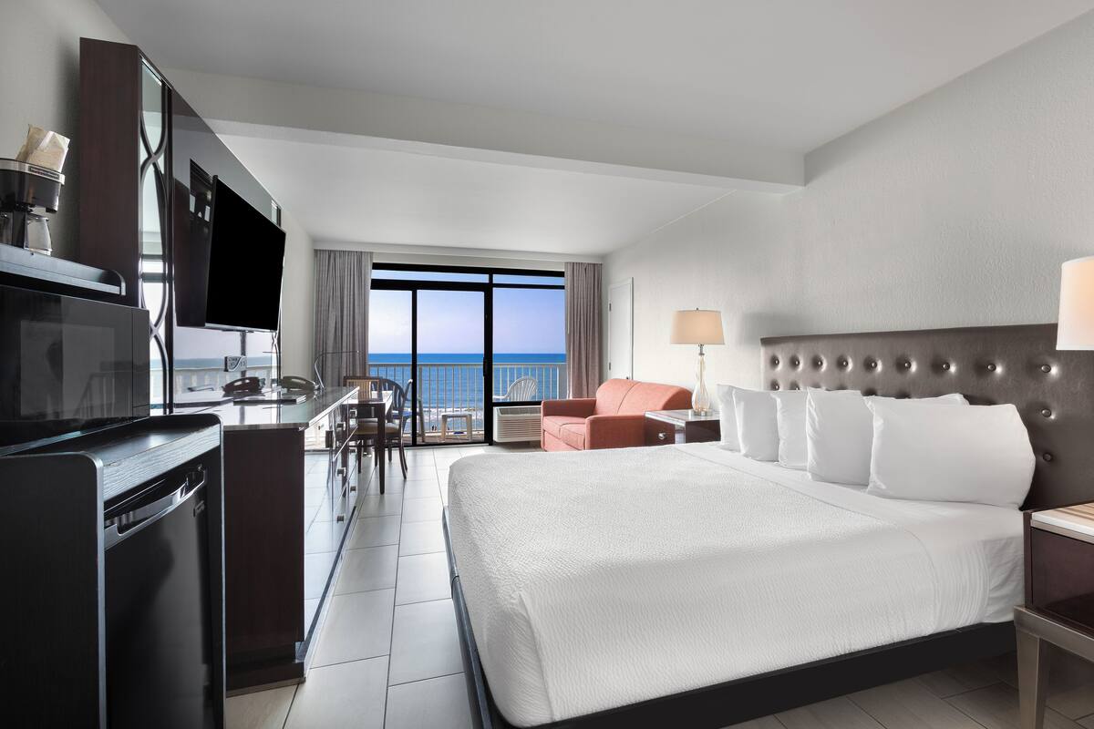 A hotel room with a large bed, a desk with a chair, a television, and a view of the ocean.
