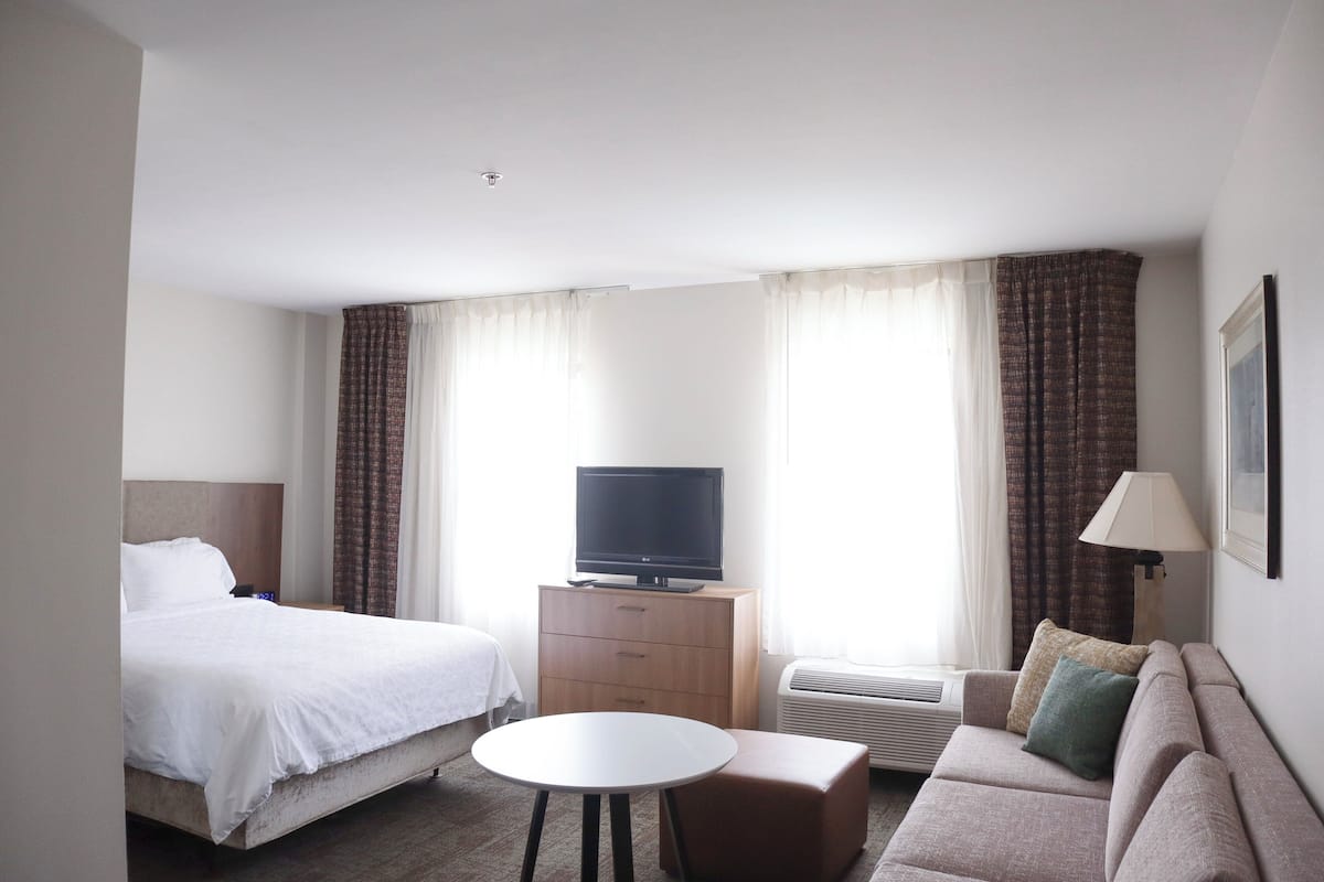 A hotel room with a bed, a TV on a wooden dresser, a sofa, a small table, and an air conditioning unit.