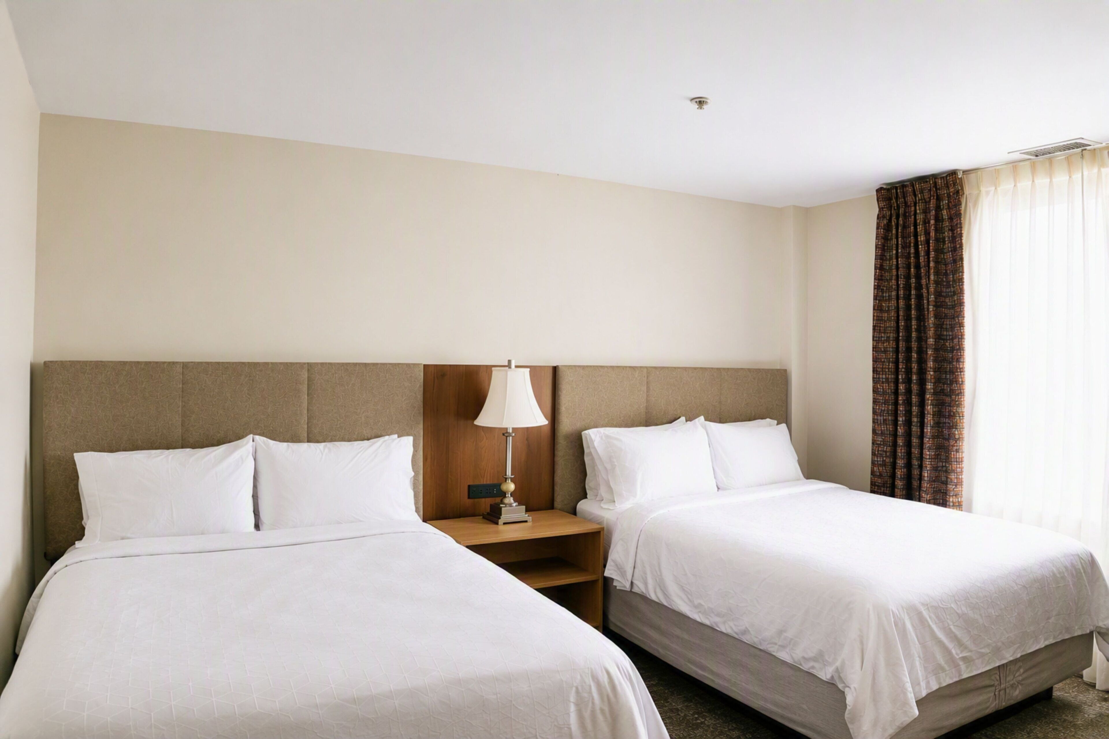A hotel room with two beds, a nightstand, a lamp, and a window with curtains.