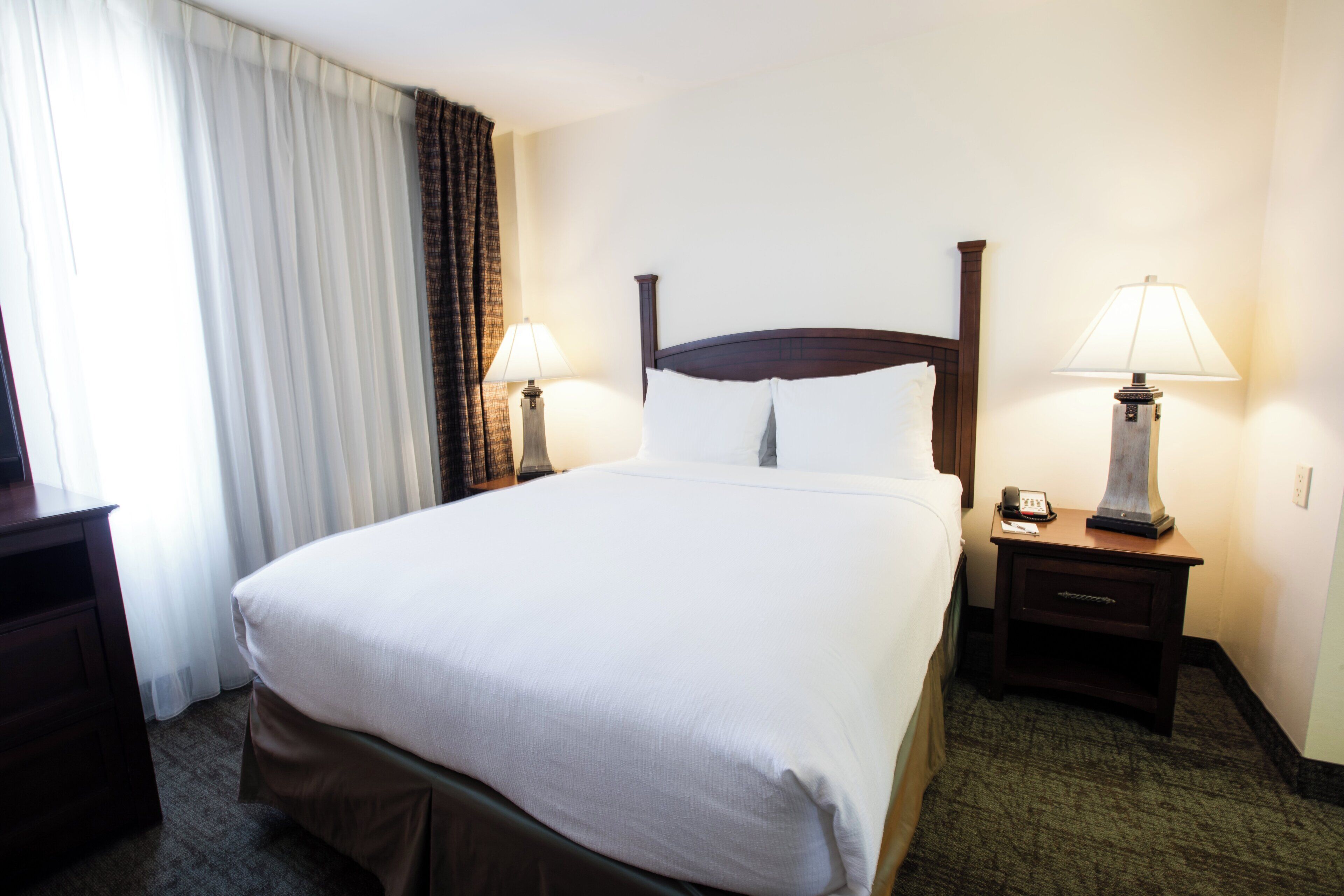 A hotel room with a large bed, two bedside tables with lamps, a nightstand, and a window with curtains.